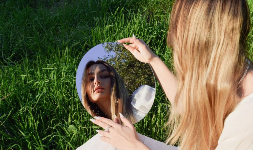 woman sitting in grass and looking in the mirror, planning to start reflection journaling routine