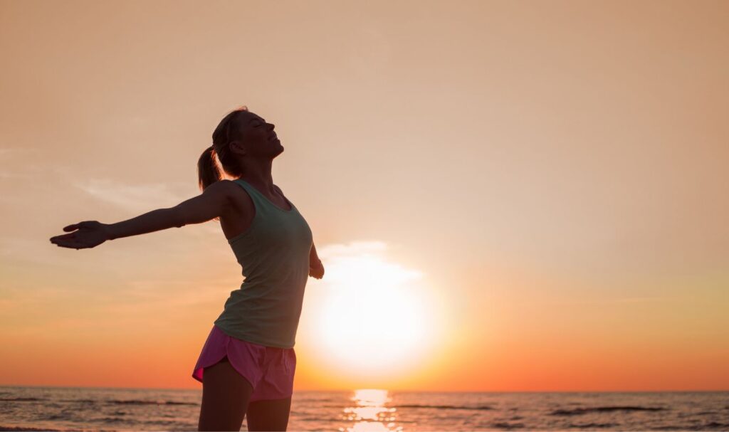 Woman standing with arms open at sunrise on the beach, symbolizing confidence, freedom, and manifesting weight loss naturally.