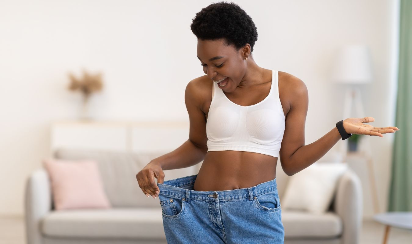 Happy woman holding oversized jeans after successful transformation, representing manifesting weight loss and healthy body confidence.