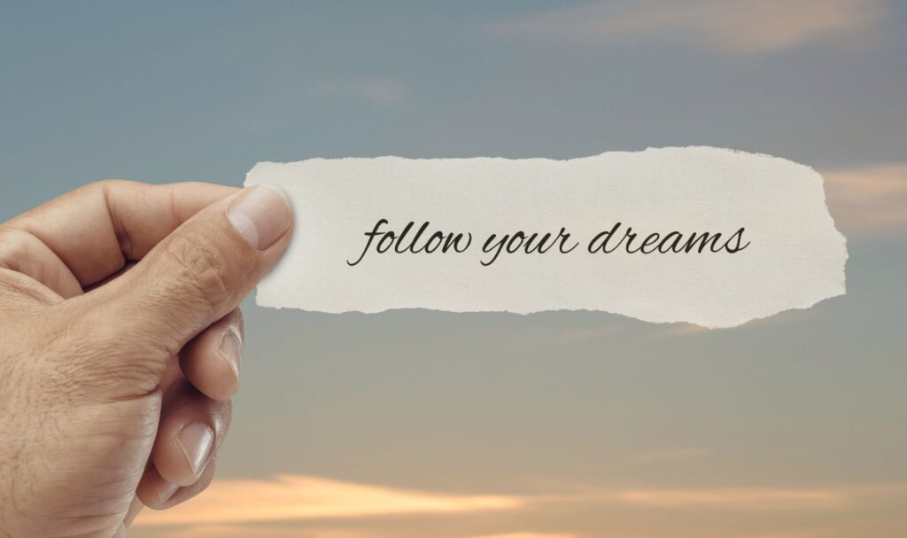 Hand holding a torn paper note reading “follow your dreams” against a soft sky background, representing motivation, intention, and manifesting your dreams into reality.