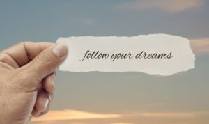 Hand holding a torn paper note reading “follow your dreams” against a soft sky background, representing motivation, intention, and manifesting your dreams into reality.