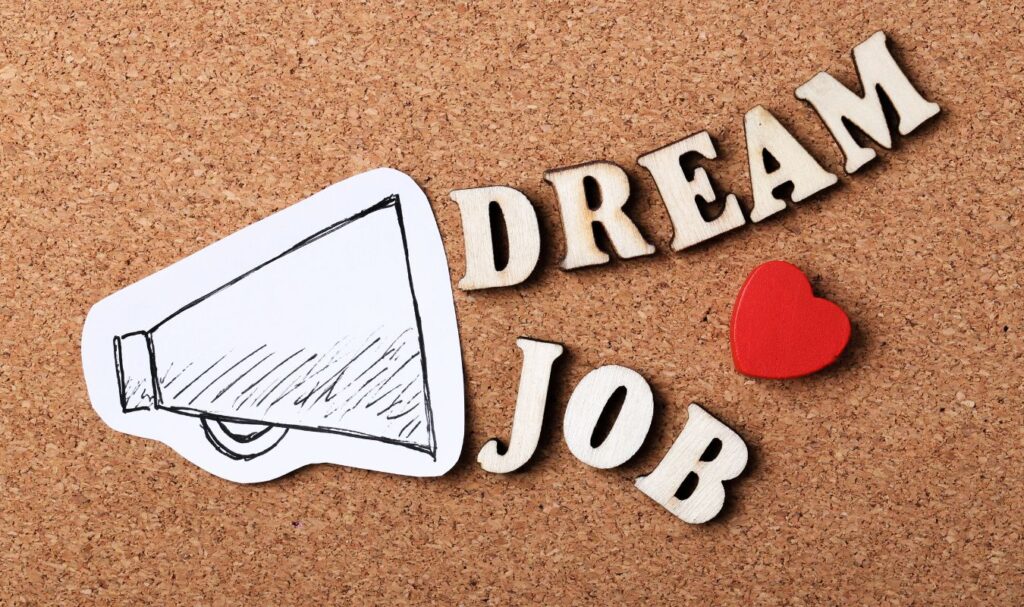 Wooden letters spelling “Dream Job” beside a megaphone and red heart on a corkboard background, symbolizing career goals, manifestation, and attracting a new job.