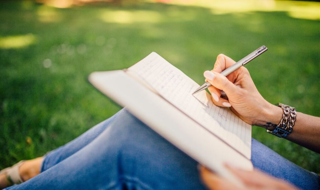 Person writing in a notebook outdoors on grass, representing journaling, intention-setting, and manifesting a new job opportunity.