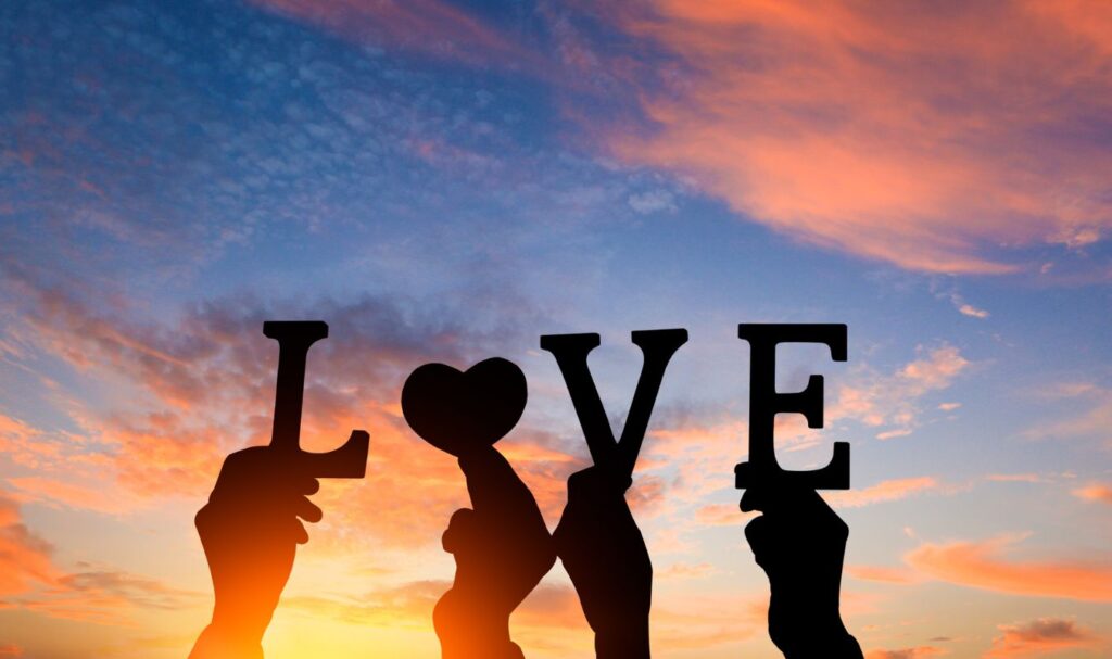 Silhouetted hands holding the word “LOVE” against a colorful sunset sky, representing romance, emotional connection, and manifesting love into your life.